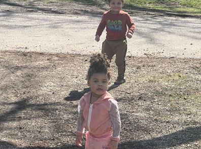 Two children walk joyfully in a sunny park, one in pink, the other in red, reflecting playful innocence and curiosity.