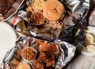 A cozy food spread with grilled meat, buns, and sides in foil, evoking warmth, comfort, and nostalgic picnic vibes.