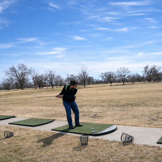 A focused golfer swings at a driving range, surrounded by grass, tees, and trees under a clear blue sky.