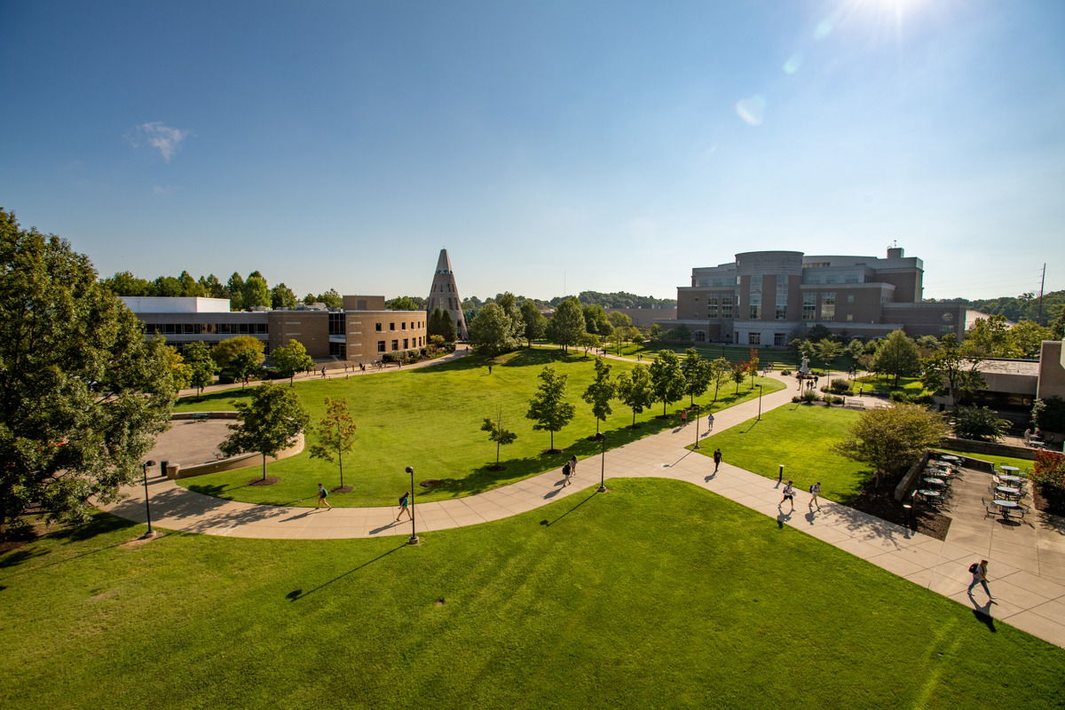 A sunny campus scene with green lawns, modern buildings, and people walking, evoking joy and a sense of community.