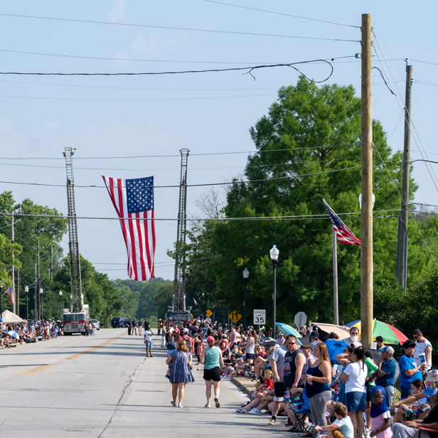 A festive street parade scene with crowds, families, American flags, and greenery, evoking excitement and community spirit.