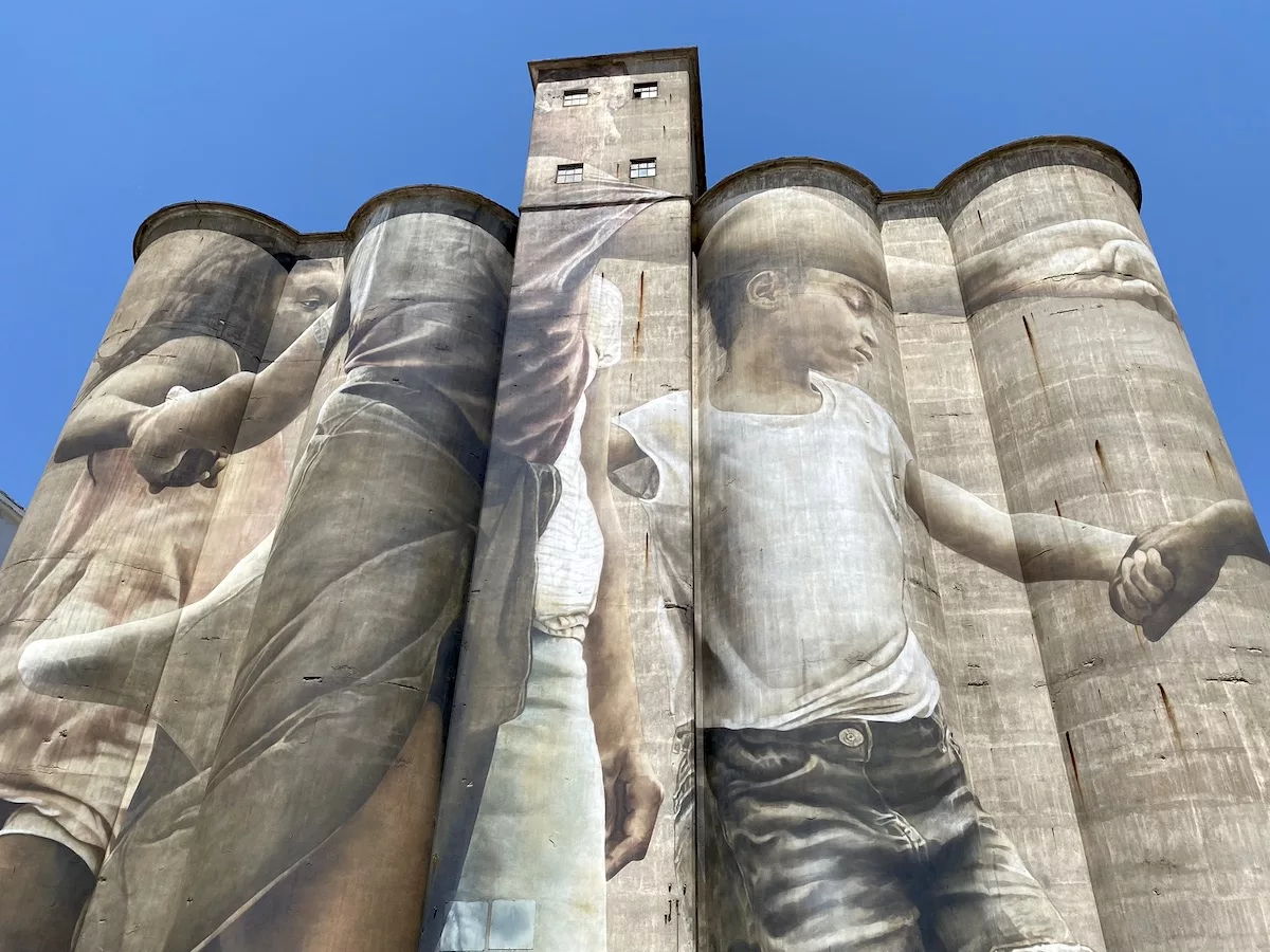 Mural on a silo shows two children holding hands, evoking feelings of innocence, nostalgia, and connection against a gray backdrop.
