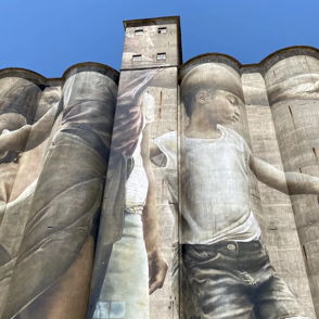 Mural on a silo shows two children holding hands, evoking feelings of innocence, nostalgia, and connection against a gray backdrop.