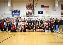Celebration of volleyball players and supporters in a gym, posing with trophies, showcasing teamwork and joy.