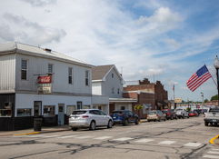 A small town street with parked cars, quaint buildings, an American flag, and a calm, nostalgic atmosphere on a pleasant day.