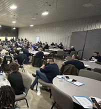Bright room with attendees at circular tables, engaged in a presentation, fostering community and collaboration.