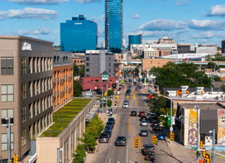 Vibrant urban landscape with tall buildings, busy street, vehicles, greenery, and a blue sky, evoking an energetic city life.