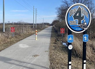 Paved Big Four Trail for pedestrians and cyclists, surrounded by sparse vegetation, power lines, and a clear blue sky.