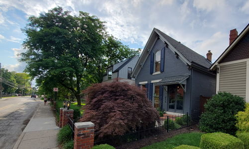 Charming street scene with two distinctive houses, lush greenery, and a partly cloudy sky, evoking tranquility and warmth.