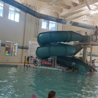 Indoor pool with turquoise slides, people swimming, and a pink float, creating a joyful, playful atmosphere for families.