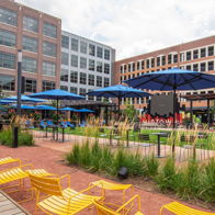 Vibrant outdoor space with modern architecture, blue umbrellas, yellow chairs, greenery, and a relaxing, inviting atmosphere.