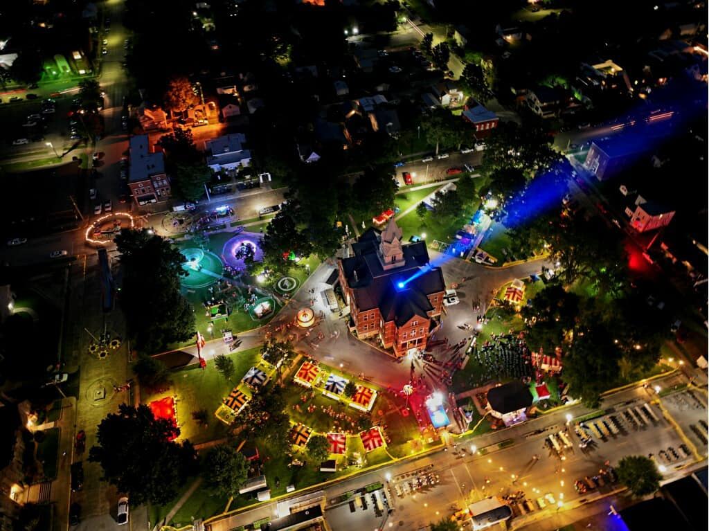 Aerial view of a vibrant nighttime festival with colorful lights, rides, and a central historic building, evoking joy and togetherness.