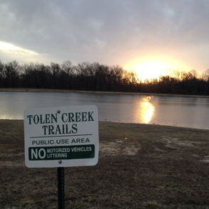 Scenic sunset view of Tolen Creek Trails, with glowing water, silhouetted trees, and a sign for public use rules.