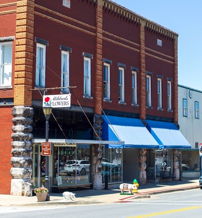 Charming small-town street with brick and painted stores, blue awnings, parked cars, and a serene atmosphere under a clear sky.