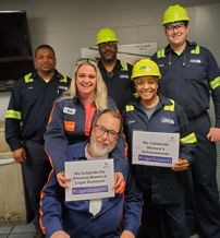 Six individuals in safety gear celebrate Women's History Month, smiling and holding signs of support for women in their field.