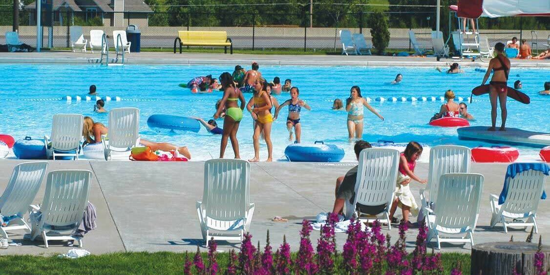 A lively pool scene with people playing, relaxing, and enjoying a sunny day among colorful inflatables and flowers.