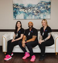 Three individuals in black scrubs relax on a couch, smiling against an abstract art backdrop, showcasing teamwork and professionalism.