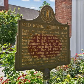 Plaque titled "Old Bank, Robbed 1868," surrounded by flowers, evokes nostalgia and appreciation for history in a serene setting.