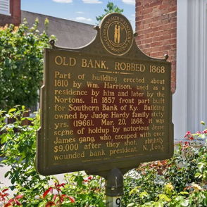 Plaque titled "Old Bank, Robbed 1868," surrounded by flowers, evokes nostalgia and appreciation for history in a serene setting.