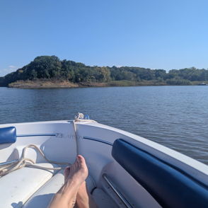 Tranquil boat scene with feet at the edge, calm water, blue sky, and greenery; evokes serenity, freedom, and nostalgia.