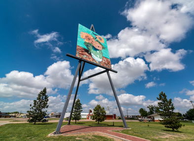 Colorful mural of sunflowers on a tall structure, bright blue sky, grass, and trees evoke a cheerful, vibrant atmosphere.