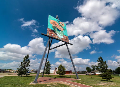 Colorful mural of sunflowers on a tall structure, bright blue sky, grass, and trees evoke a cheerful, vibrant atmosphere.