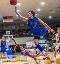 Basketball player in blue jersey leaps for a shot, challenged by an opponent, showcasing athleticism and competitive intensity.