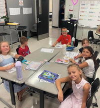A cheerful group of children collaborates at a classroom table, surrounded by notebooks and colorful materials, enjoying learning.