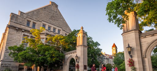 A vibrant campus scene at sunset, featuring a stone building, blooming flowers, and people enjoying a welcoming atmosphere.