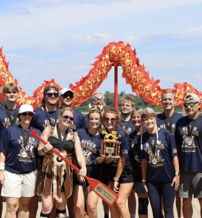 Group of smiling people in matching shirts on a sunny beach, celebrating together with a colorful dragon decoration in the background.