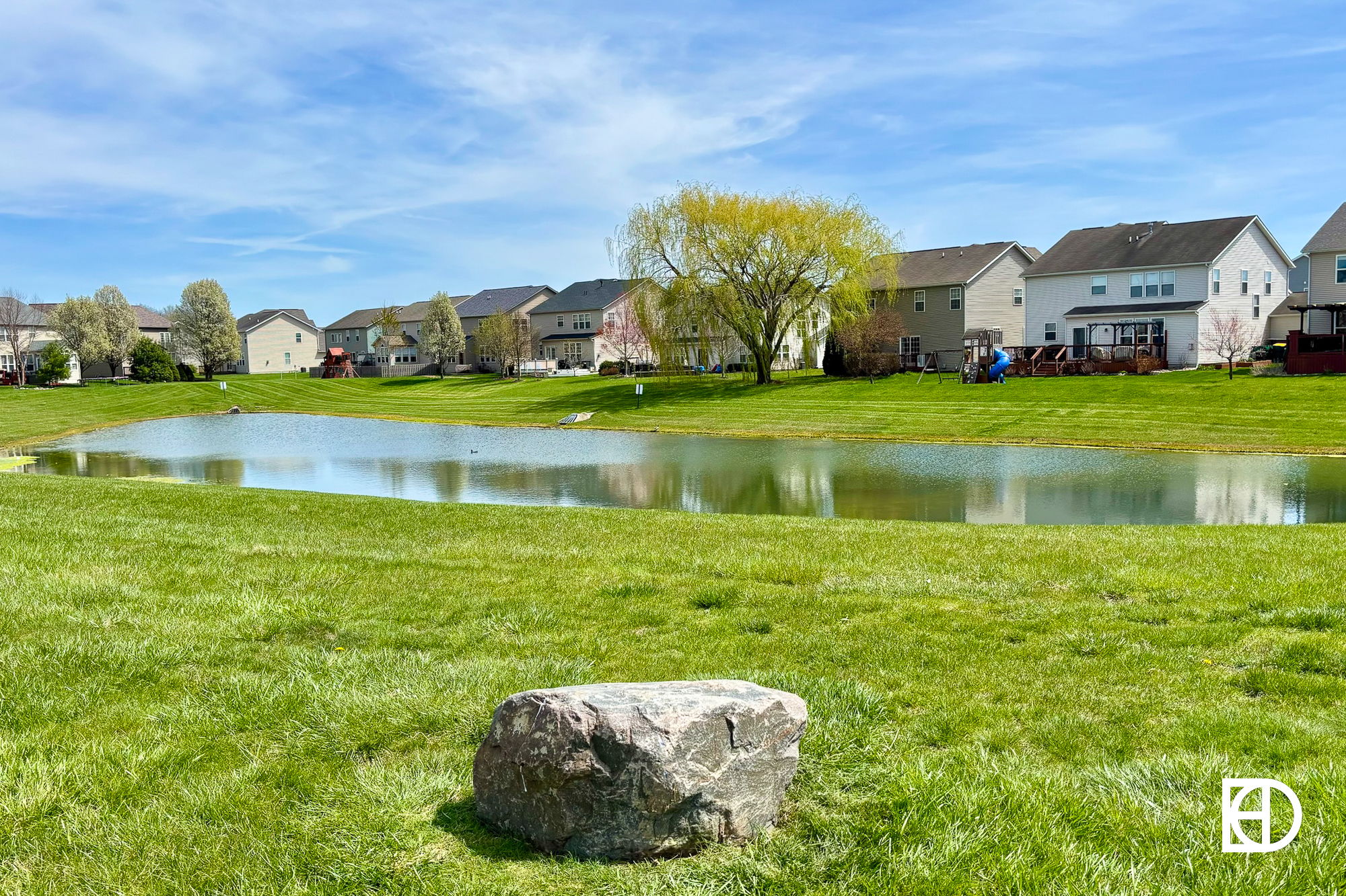 Serene landscape with a lush lawn, calm pond, houses, and a rock under a clear blue sky, evoking tranquility and peace.