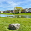 Serene landscape with a lush lawn, calm pond, houses, and a rock under a clear blue sky, evoking tranquility and peace.