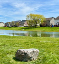 Serene landscape with a lush lawn, calm pond, houses, and a rock under a clear blue sky, evoking tranquility and peace.