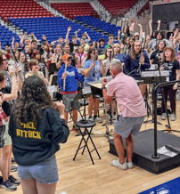 A lively ensemble of diverse musicians rehearses enthusiastically under a conductor, showcasing unity and passion for music.