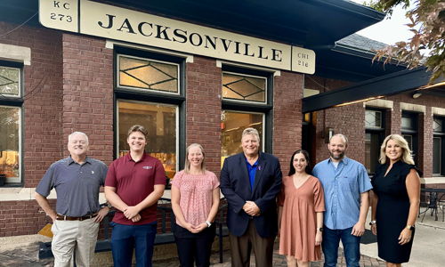 Group of seven people smiling outside a lit building with a "JACKSONVILLE" sign, exuding warmth and camaraderie.