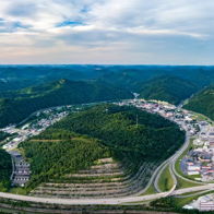 Panoramic view of lush mountains, green hills, highways, and a quaint town under a partly cloudy sky, evoking tranquility and wanderlust.