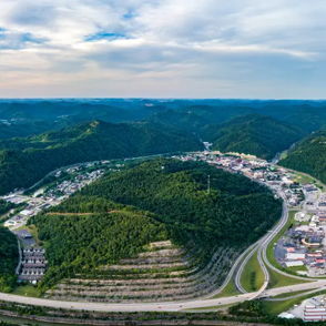 Panoramic view of lush mountains, green hills, highways, and a quaint town under a partly cloudy sky, evoking tranquility and wanderlust.