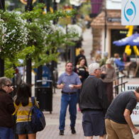 Busy urban street scene with diverse people, shops, greenery, and cheerful decorations, evoking community and connection.