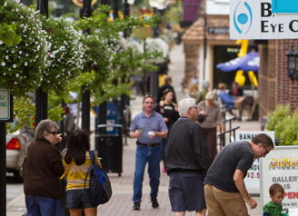 Busy urban street scene with diverse people, shops, greenery, and cheerful decorations, evoking community and connection.