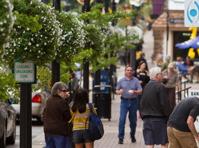 Busy urban street scene with diverse people, shops, greenery, and cheerful decorations, evoking community and connection.