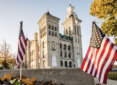 Historic building with towers, small flags in foreground, autumn leaves, evoking patriotism, warmth, and community pride.