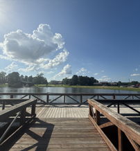 Serene lakeside scene with a wooden dock, calm water, bright blue sky, and fluffy clouds, evoking tranquility and relaxation.