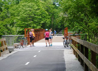 Outdoor scene with cyclists, pedestrians, and people socializing near a bridge in a bright, lush, and inviting setting.