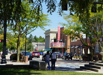 Lively urban street scene with figures conversing, lush trees, a theater backdrop, vehicles, and a sunny, cheerful atmosphere.