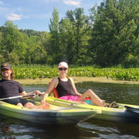 Two people in kayaks on calm water, surrounded by greenery, smiling in the sun, evoking joy and tranquility.