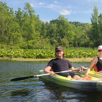 Two people in kayaks on calm water, surrounded by greenery, smiling in the sun, evoking joy and tranquility.