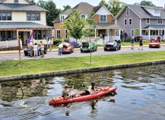 Two people kayak in calm waters by charming homes; a group onshore enjoys a sunny day, enhancing the scene's warmth and community.