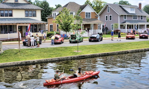 Two people kayak in calm waters by charming homes; a group onshore enjoys a sunny day, enhancing the scene's warmth and community.