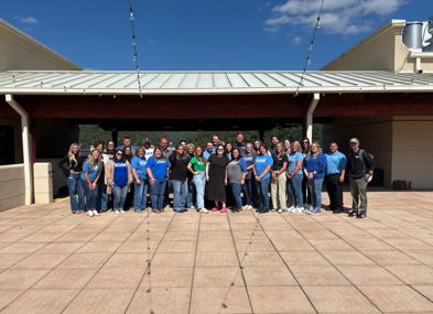 A joyful outdoor gathering with people in matching blue shirts, celebrating teamwork under a sunny sky.