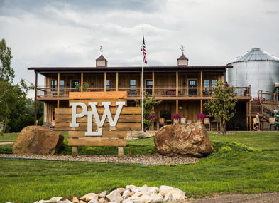 Rustic building with "PLW" sign, wooden facade, stone accents, surrounded by greenery and silos, evoking warmth and tranquility.